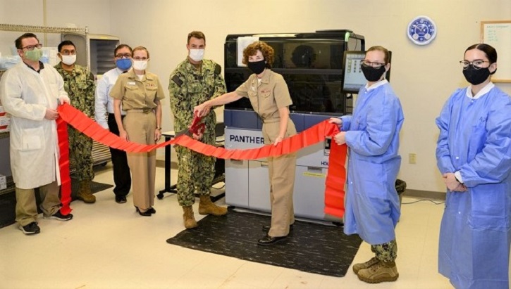 Image of A group of military health personnel, wearing masks, in front of a large machine with a red ribbon in front of it. .