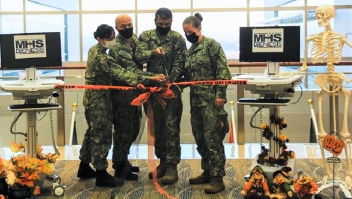 Image of Four military personnel, wearing masks, standing in front of two computer screens that say "MHS GENESIS.".
