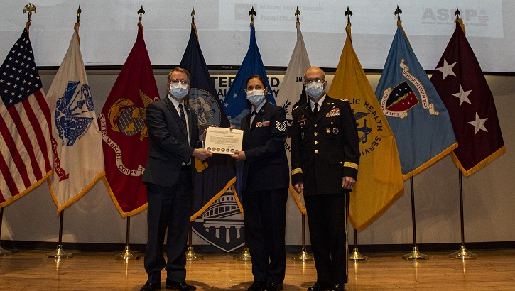 Image of Three military personnel in uniform, wearing masks, in front of flags. Click to open a larger version of the image.