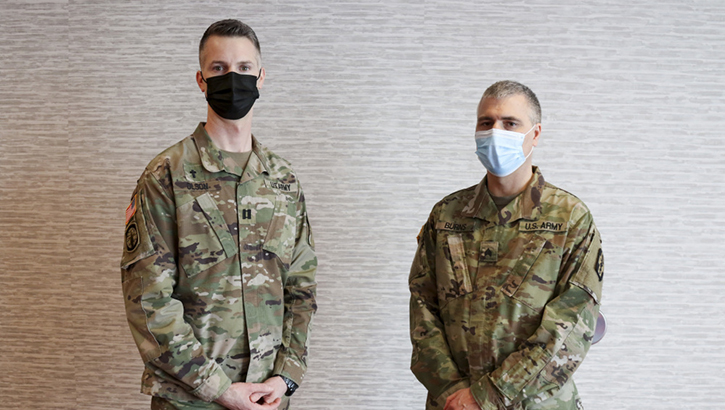 Image of Two military personnel, wearing masks, standing against a wall.