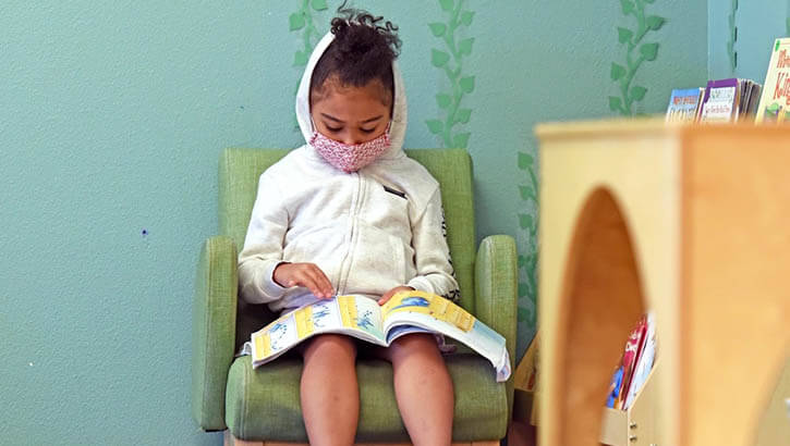 Image of 5-year-old girl in mask reads a book by herself.