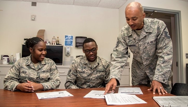 Three soldiers at a desk, two sitting and one standing, pointing at a piece of paper