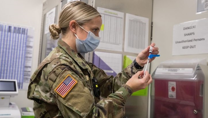 A military nurse, wearing a mask, prepares a needle for a vaccination
