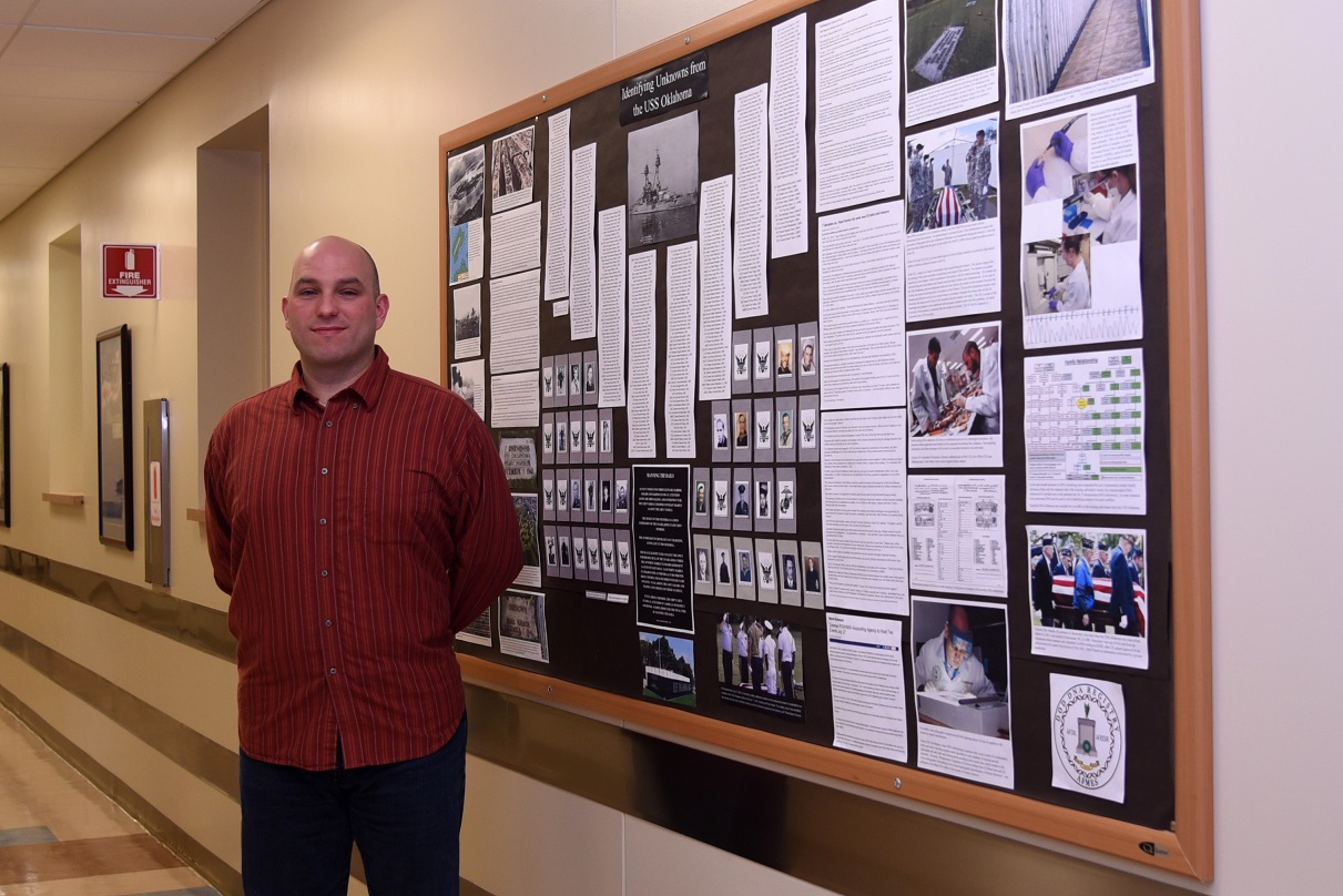 Sean Patterson, Armed Forces Medical Examiner System Department of Defense DNA Registry DNA analyst, stands in front of the USS Oklahoma History Board Nov. 29, 2016, at AFMES on Dover Air Force Base, Delaware. The board tells what happened to the USS Oklahoma during the attack on Pearl Harbor and the difficulty of identifying the 429 service members who were lost. (U.S. Air Force photo/Senior Airman Ashlin Federick)