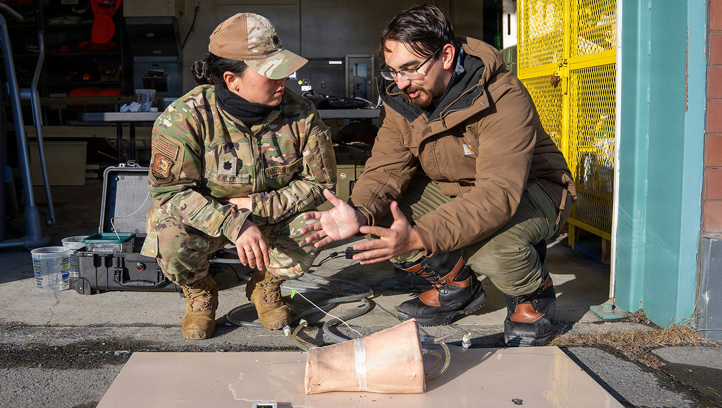 Andres Martinez Murillo, lead biomedical engineer for Naval Medical Research Unit (NAMRU) San Antonio, explains stability testing on the novel SynDaver thermal, bleeding manikin thigh to U.S. Air Force Lt. Col. Kaitlin Salle at the Kodiak Coast Guard Clinic