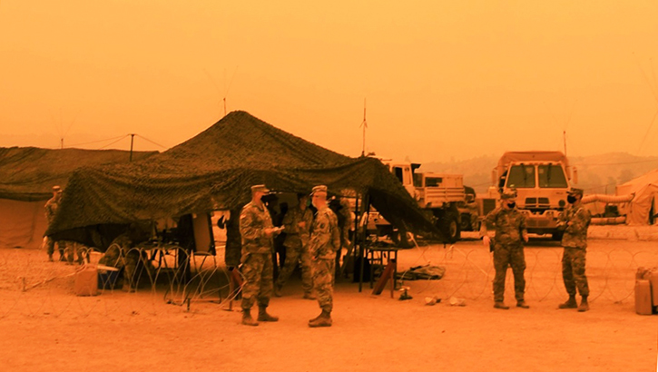 Picture of a military tent; an orange, smoky hue surrounds the tent and soldiers