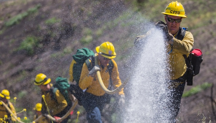Men in protective suits dousing a flame with water from a hose