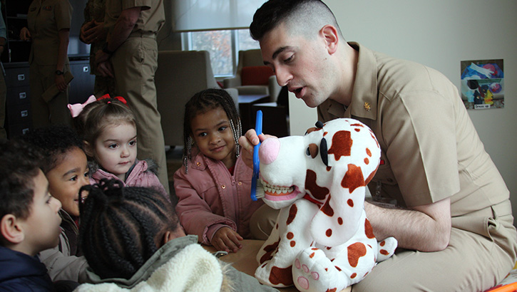 Image of Military health personnel, sitting in front of a group of children, showing them how to brush their teeth using a stuffed animal.