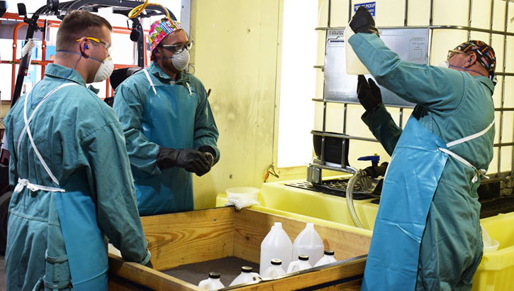 Image of three men in PPE examining bottles of hand sanitizer. Click to open a larger version of the image.