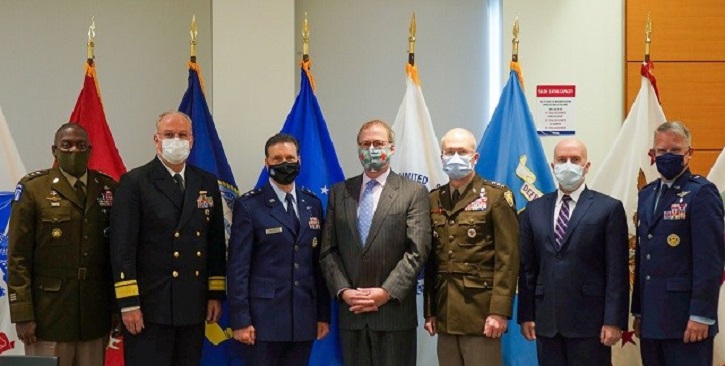 Image of Military personnel, wearing masks, standing in a line in front of flags. Click to open a larger version of the image.