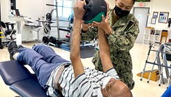 A female Navy physical therapist works with a senior citizen lying on a table holding a ball.