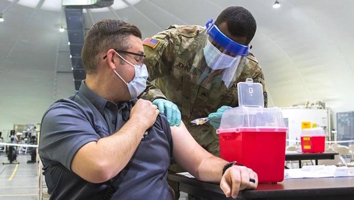 Image of Medical director at Fort Riley, Kansas receives a COVID-19 vaccination In his left arm from a tech in personal protective equipment.