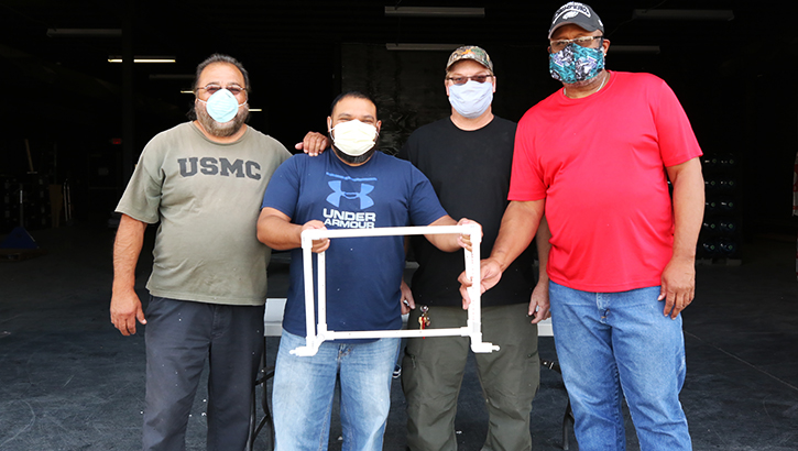 Image of Four men wearing masks, holding COVID-19 Airway Management Isolation Chamber. Click to open a larger version of the image.