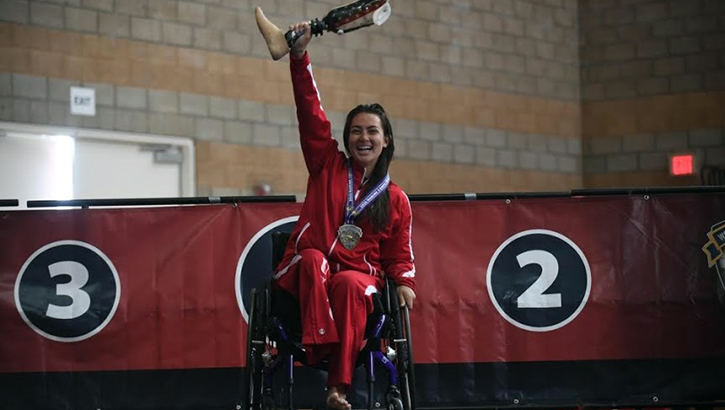 Woman in wheelchair on podium; holding up prosthetic leg