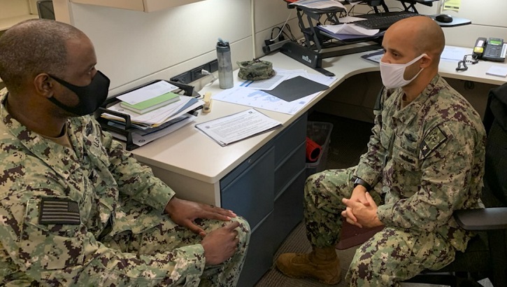 Two military personnel, wearing masks, sitting at a desk talking