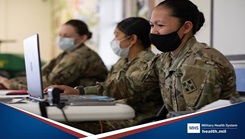 Three military personnel, wearing masks, sitting at a desk and typing on laptops 