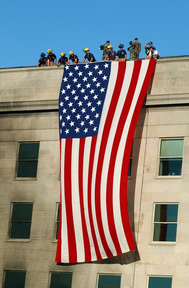 Military Service members render honors as fire and rescue workers unfurl a huge American flag over the side of the Pentagon during rescue and recovery efforts following the Sept 11 terrorist attack. The attack came at approximately 9:40 a.m. as a hijacked commercial airliner, originating from Washington D.C.'s Dulles airport, was flown into the southern side of the building facing Route 27. (U.S. Navy photo by Photographer's Mate 1st Class Michael W. Pendergrass)