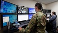 Uniformed service member stands behind wall of computer screens 