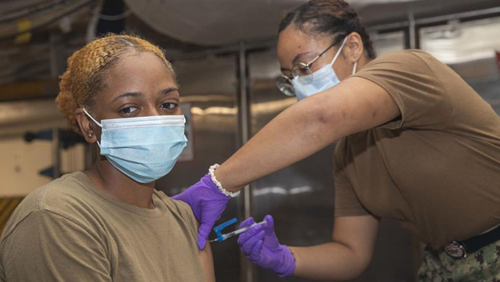 Female hospital corpsman gives a COVID-19 vaccine injection to a sailor in her left arm