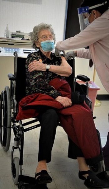 A lady in a wheel chair wearing a face mask receives the COVID-19 vaccine from military health personnel