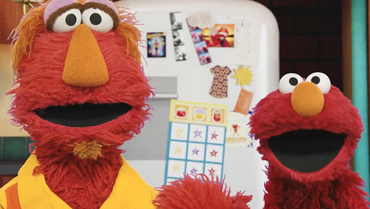 A video still shows the Muppet Elmo and his father looking toward the camera.
