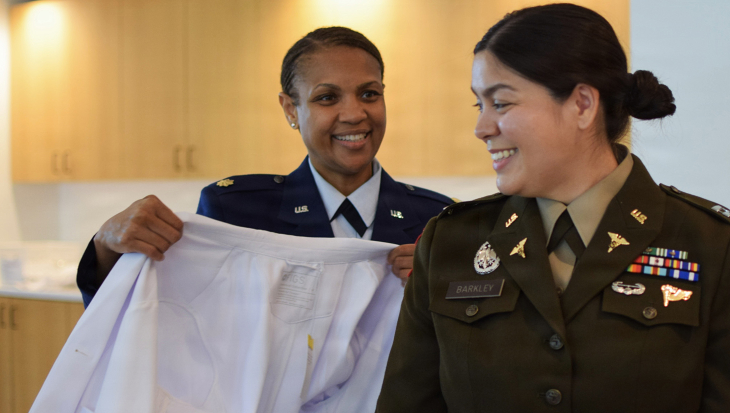 U.S. Air Force Maj. Barbara Jean (left), site director for the Interservice Physician Assistant Program Phase 2 at Walter Reed National Military Medical Center, assists U.S. Army Capt. Bonnie Claire Barkley don her white coat