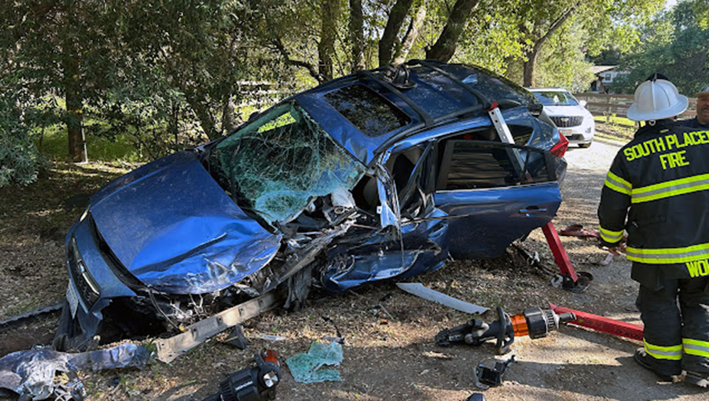 A vehicle rests heavily damaged against the tree line following a nearly head-on collision